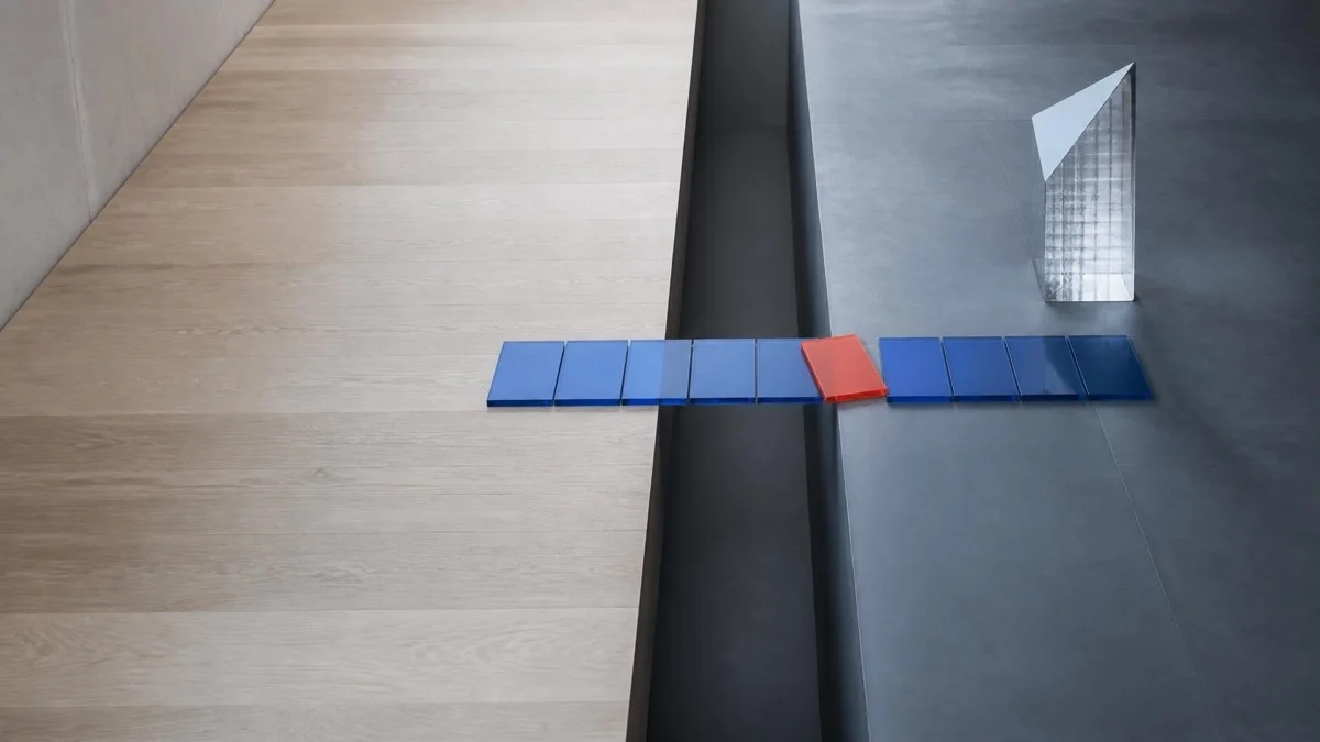 Overhead view of a row of blue glass tiles with one red tile crossing a threshold between wood and concrete floors, symbolizing 360 feedback across competencies