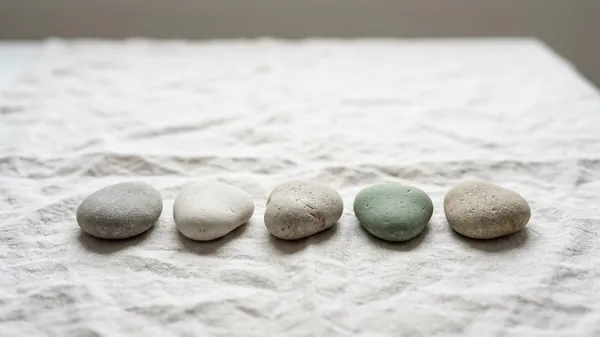 Five smooth stones in a row on natural linen, four in grey and cream tones and one sage green, illustrating the 5 qualities of a good leader