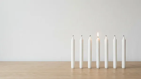 Seven white taper candles standing in a row on a pale oak table against a white wall, one of them lit, illustrating 7 leadership traits you can watch someone do