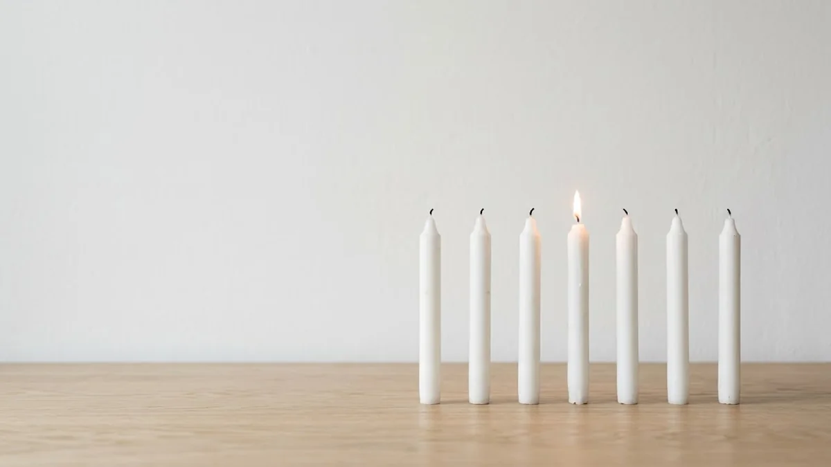 Seven white taper candles standing in a row on a pale oak table against a white wall, one of them lit, illustrating 7 leadership traits you can watch someone do