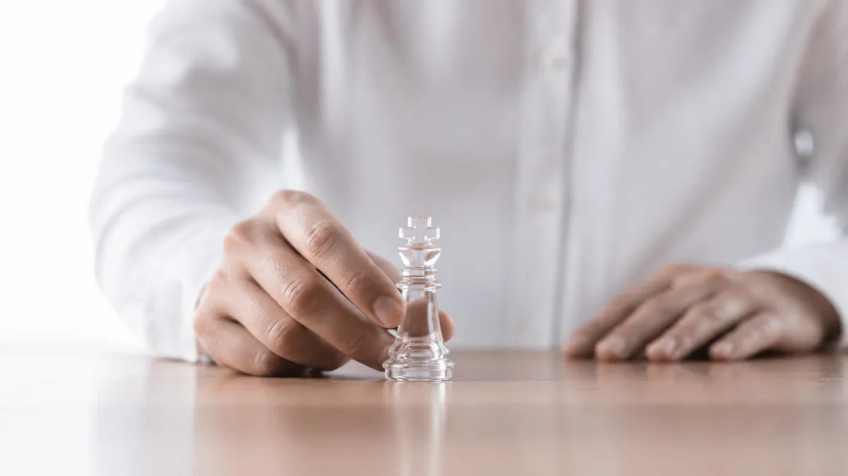 A hand carefully moving a clear glass king chess piece on a table, symbolizing the strategic moves that keep AI leadership rollouts from stalling