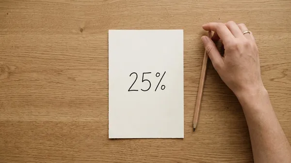Overhead view of a sheet of cream paper with 25% handwritten on it on an oak desk, a hand resting on a pencil beside it, illustrating the 25% Rule for a check-in meeting