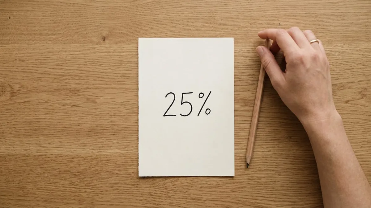 Overhead view of a sheet of cream paper with 25% handwritten on it on an oak desk, a hand resting on a pencil beside it, illustrating the 25% Rule for a check-in meeting