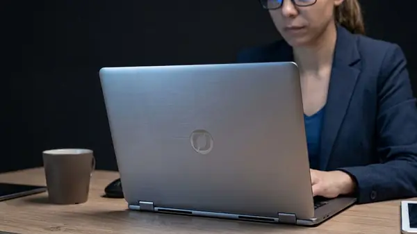 A woman in glasses and a navy blazer typing on a silver laptop at a wooden desk beside a mug and a tablet, illustrating a CEO writing a chief of staff job description