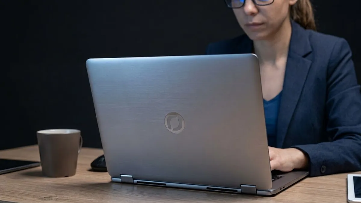 A woman in glasses and a navy blazer typing on a silver laptop at a wooden desk beside a mug and a tablet, illustrating a CEO writing a chief of staff job description