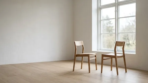 Two oak chairs angled toward each other beside a tall window in an empty white room with a pale wood floor, illustrating the setting for a difficult conversation between a manager and an employee