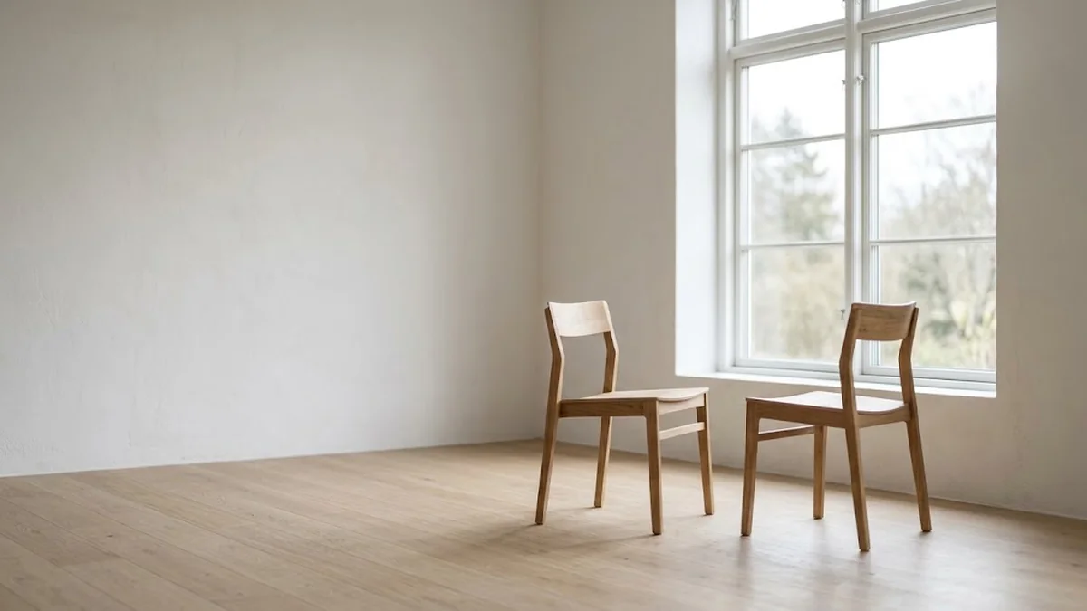 Two oak chairs angled toward each other beside a tall window in an empty white room with a pale wood floor, illustrating the setting for a difficult conversation between a manager and an employee