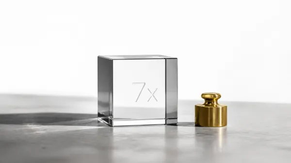 A clear glass cube laser-etched with '7x' beside a small brass weight on a concrete surface, symbolizing the measured return on investment from executive coaching