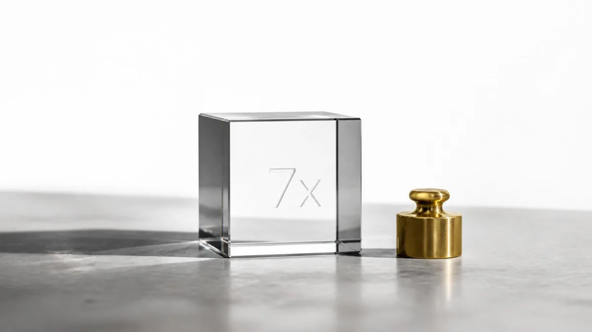 A clear glass cube laser-etched with '7x' beside a small brass weight on a concrete surface, symbolizing the measured return on investment from executive coaching