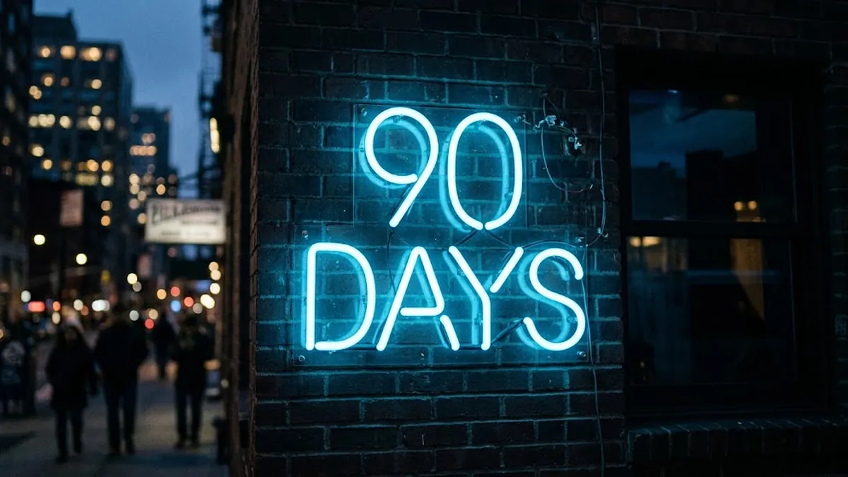 A blue neon sign reading 90 Days on a dark brick wall beside a city street at dusk, illustrating the first 90 days of a first-time manager leading people