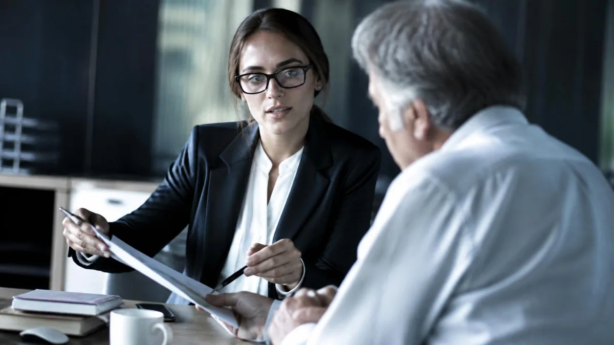 Professional woman making her case to her manager across a desk with a one-page document, on getting your company to pay for executive coaching