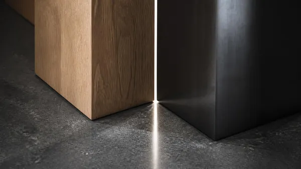 A solid oak block and a dark steel block standing side by side on a stone floor with a thin line of light passing through the narrow gap between them, illustrating the handoff point in how to delegate so the work stays delegated