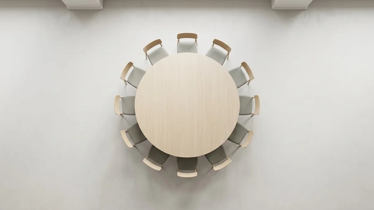 Overhead view of a round pale oak meeting table ringed by matching chairs in a bright empty room, illustrating the weekly Level 10 Meeting of a leadership team