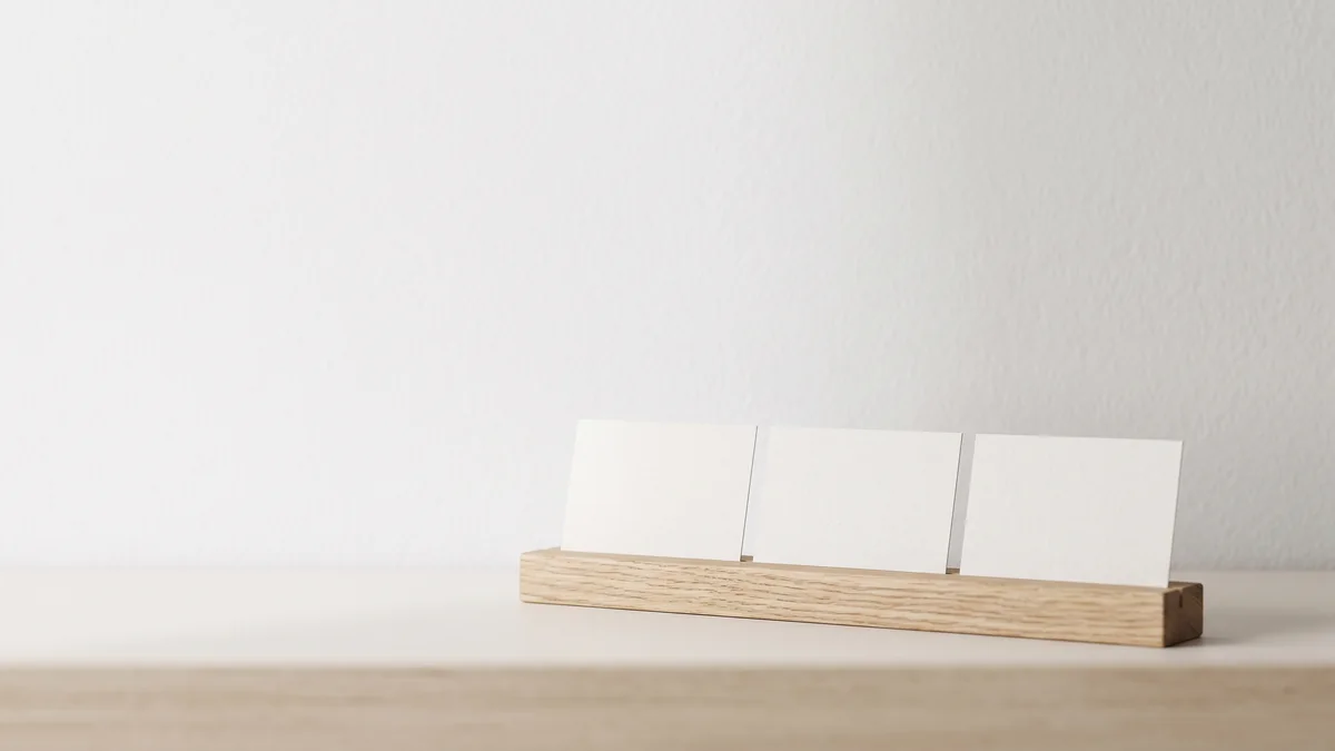 Three blank white cards standing side by side in a pale oak holder on a shelf against a white wall, illustrating performance improvement plan examples laid out for comparison