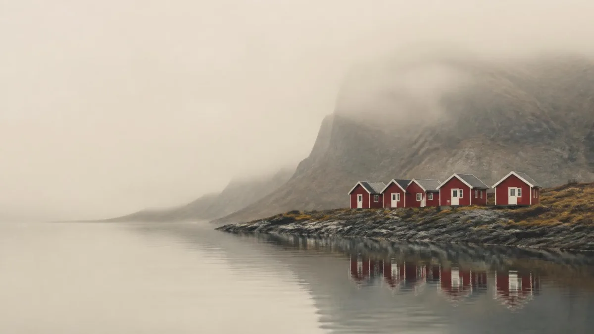 Row of identical red wooden cabins along a misty fjord shore beneath a mountain, evoking the calm egalitarian Scandinavian leadership style