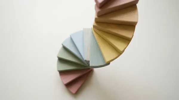 A spiral staircase of pastel wooden wedges in pink, sage, blue, yellow and tan seen from above on a white surface, illustrating the skip-level meeting as a way for leaders to hear from every level