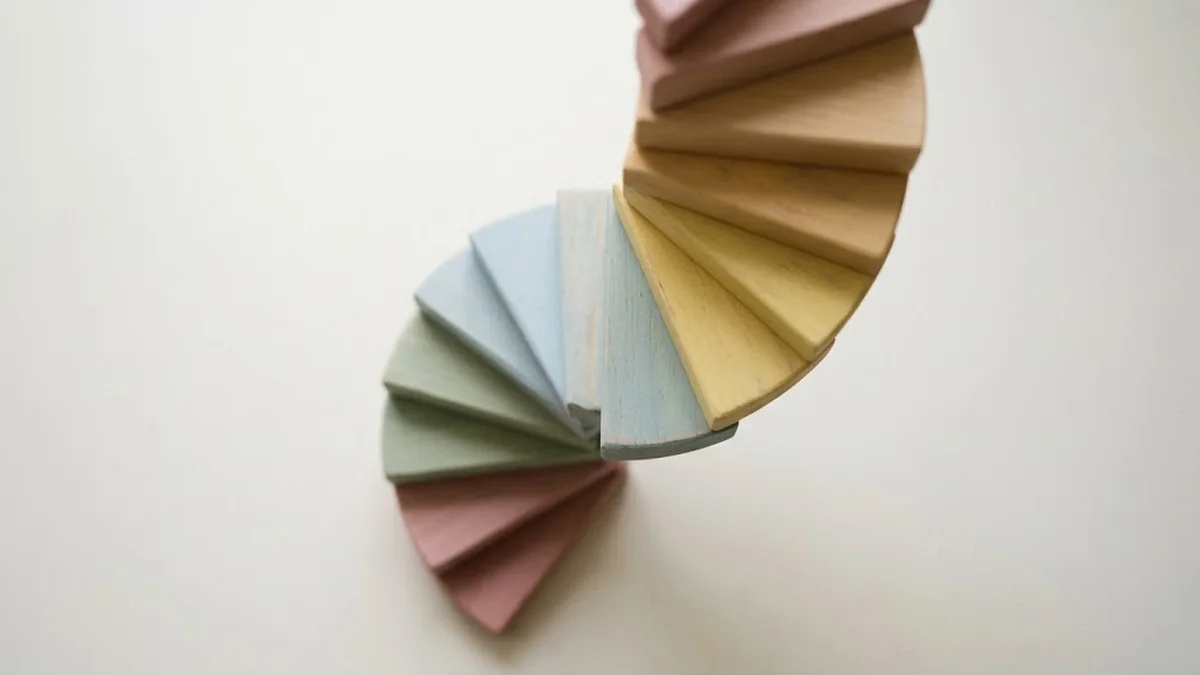 A spiral staircase of pastel wooden wedges in pink, sage, blue, yellow and tan seen from above on a white surface, illustrating the skip-level meeting as a way for leaders to hear from every level