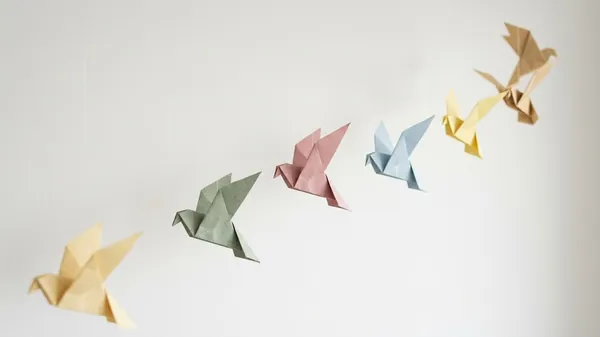 Six origami paper birds in pastel yellow, sage, pink, blue and tan hanging in a rising diagonal line against a white wall, illustrating the types of followership and the different followers a company produces